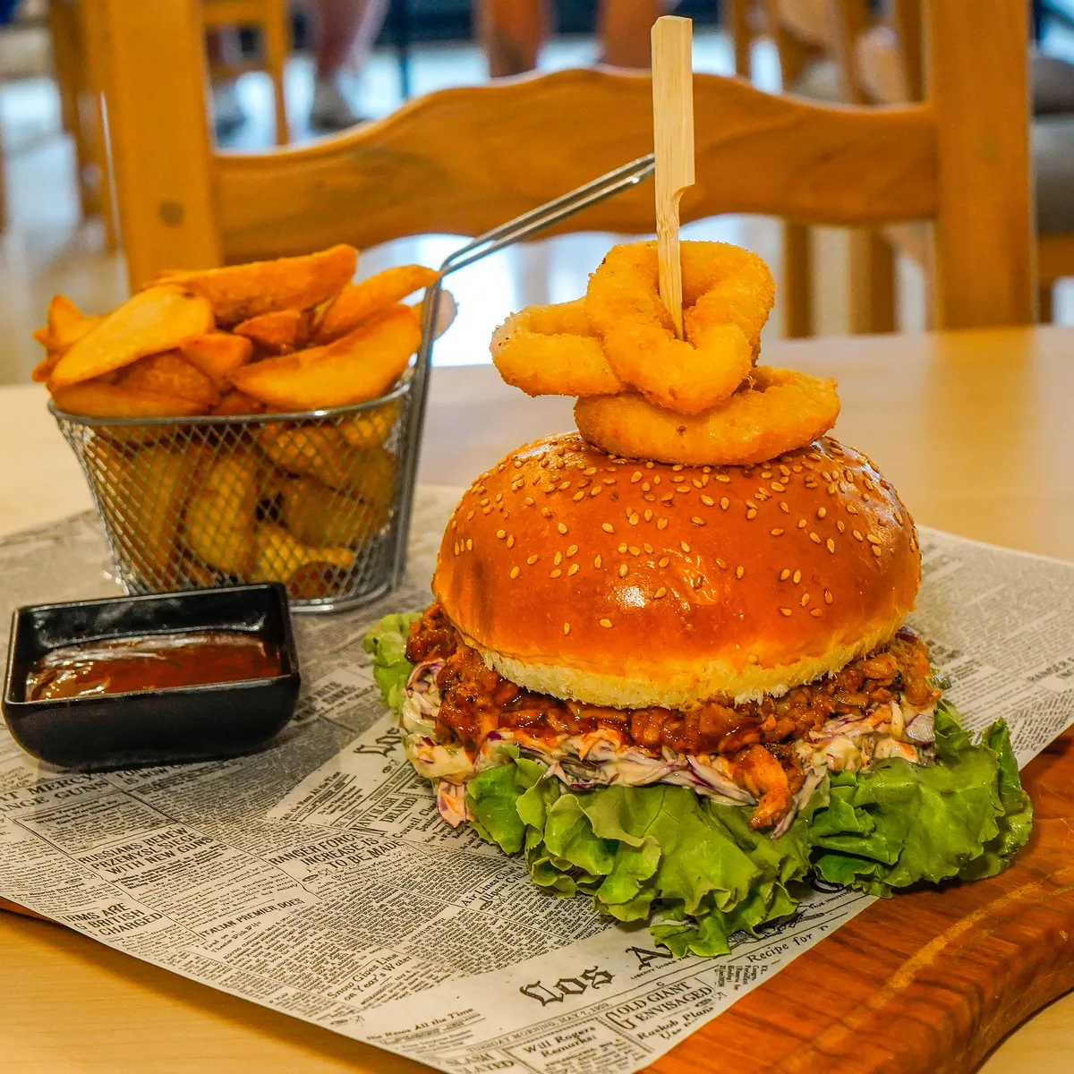 Burger s čebulnimi obročki, krompirjevimi krhlji v košarici in omako na leseni deski.